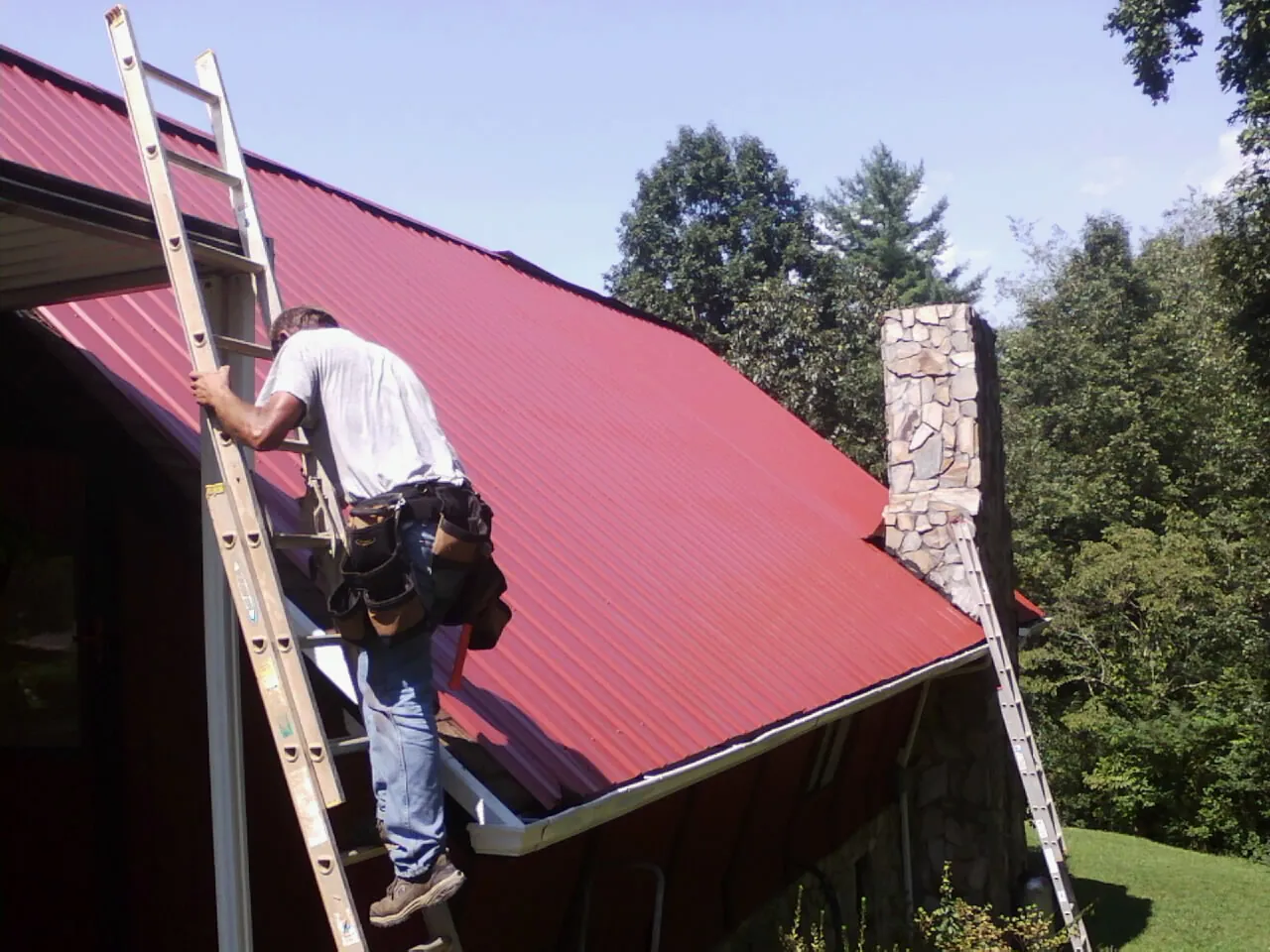 Roofer on a ladder beside a red metal roof and stone chimney — Soffit & Fascia Repair by Roofing Brothers in Crompond
